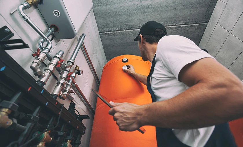 Service technician examining gauge on an orange heating appliance in a boiler room.